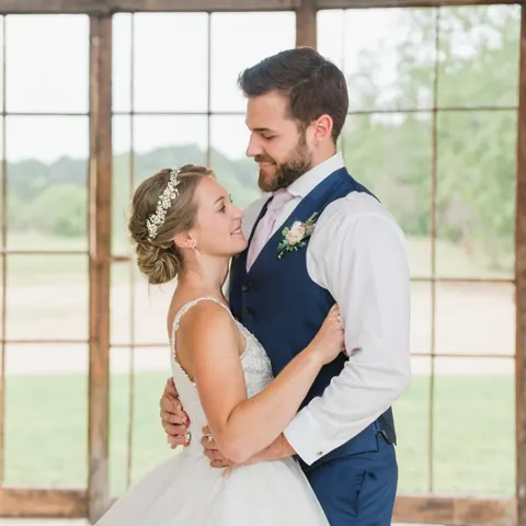 A bride and groom embrace warmly in front of a rustic window overlooking a green field.