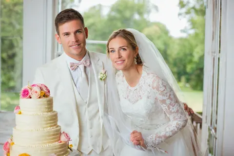 A bride and groom smiling beside a decorated wedding cake near a window.