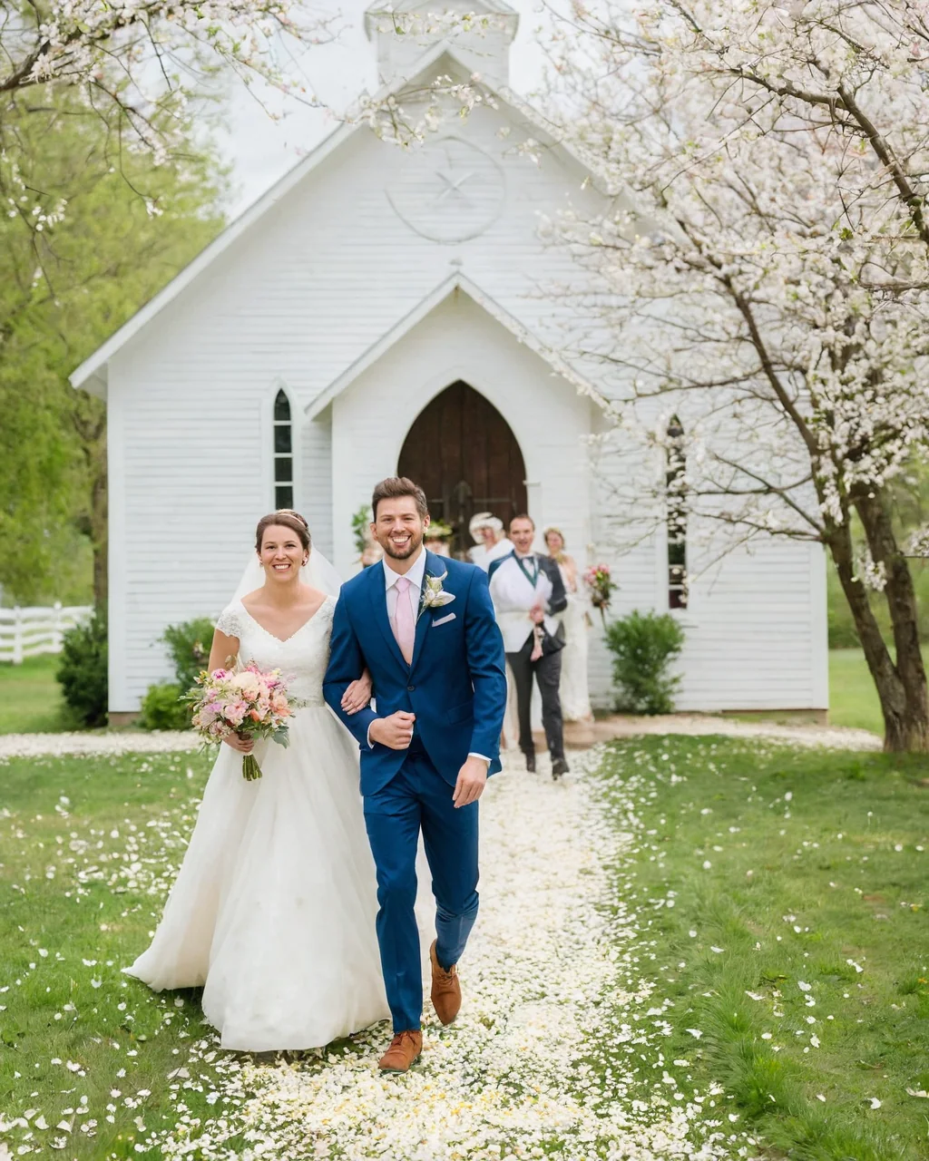 A bride and groom walk down a petal-covered path toward a white chapel surrounded by cherry blossoms.