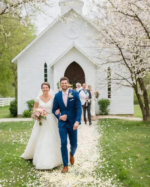 A bride and groom walk down a petal-covered path toward a white chapel surrounded by cherry blossoms.