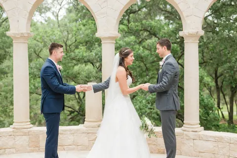 A bride in a white gown stands between two grooms holding hands under a stone arbor in a green garden.