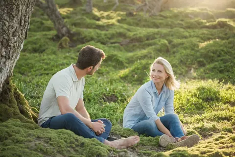 A man and woman sitting on mossy ground under a tree smiling at each other.