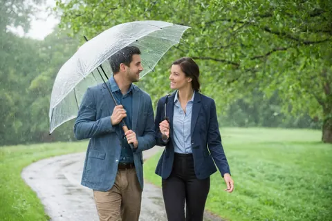 A man and woman smiling while walking under an umbrella in the rain on a park path.