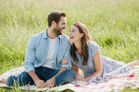 A smiling couple laughing together on a blanket in a grassy field.