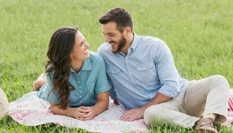 A smiling couple sitting on a floral blanket in a grassy field.