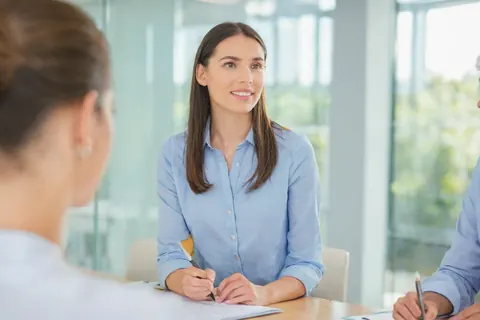 A woman in a blue shirt smiles while participating in a business meeting at a conference table.