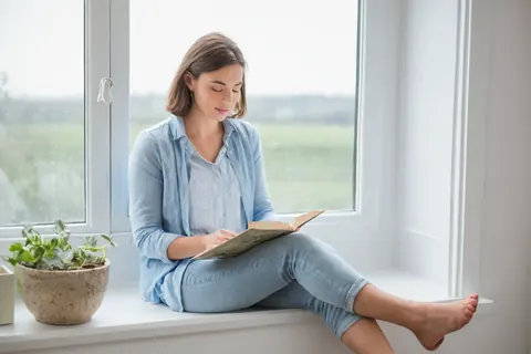 A woman in blue clothes reads a book on a windowsill next to a potted plant.