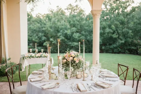 Elegant outdoor dining table set with candles flowers and glassware under a colonnade overlooking a garden.