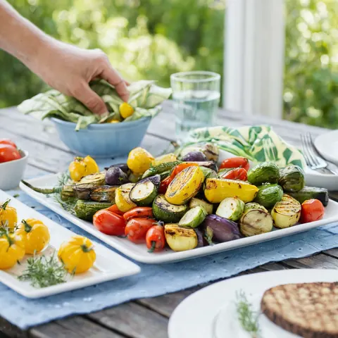 Grilled vegetables and cherry tomatoes on a wooden table with a hand reaching for a bowl.