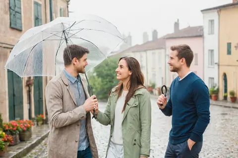 Three people smiling and chatting under an umbrella on a rainy cobblestone street.