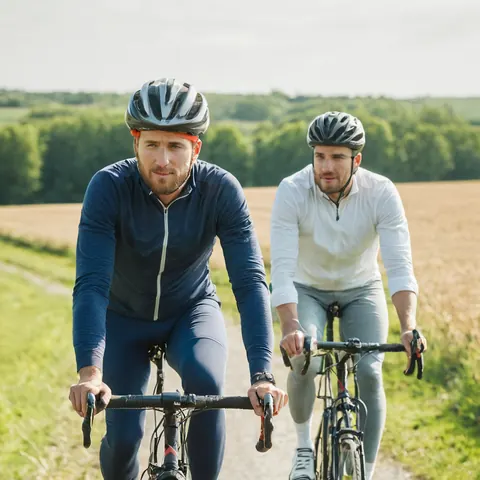Two men cycling on a rural road through golden fields and green trees on a sunny day.