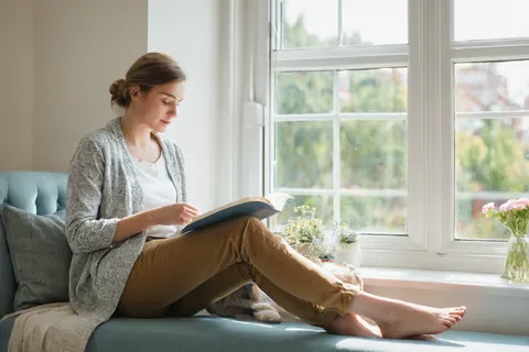 Woman reading a book on a blue sofa near a sunny window with flowers outside.